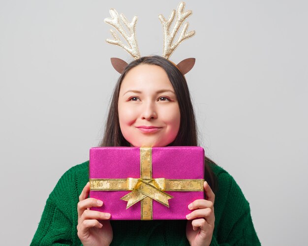 Cute young funny woman dressed as a christmas deer rejoices with a gift on a light background