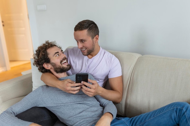 Cute young gay couple video calling their friends in their\
living room at home two male lovers smiling cheerfully while\
greeting their friends on a smartphone young gay couple sitting\
together