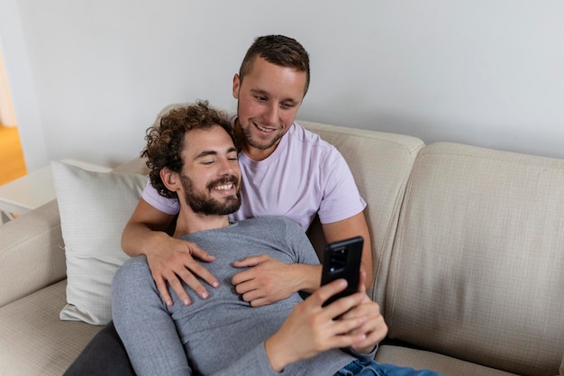 Photo cute young gay couple video calling their friends in their living room at home two male lovers smiling cheerfully while greeting their friends on a smartphone young gay couple sitting together