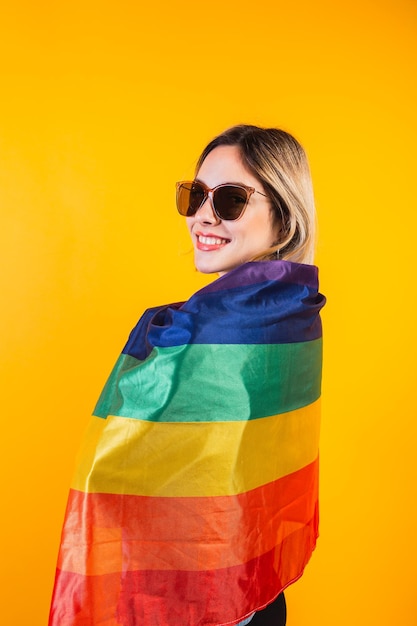 Photo cute young girl drapes herself with large rainbow flag on yellow background.