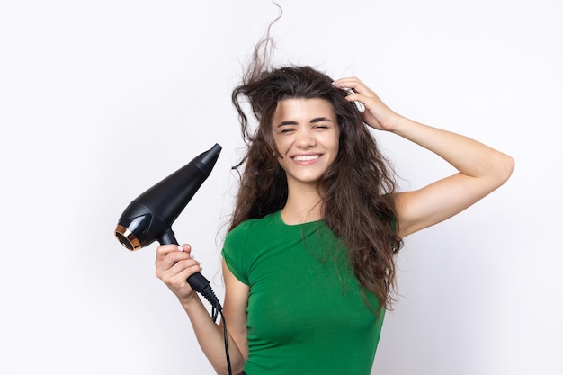 Foto una giovane ragazza carina vestita con un top verde asciuga i suoi bellissimi capelli lunghi e setosi con un asciugacapelli su uno sfondo bianco