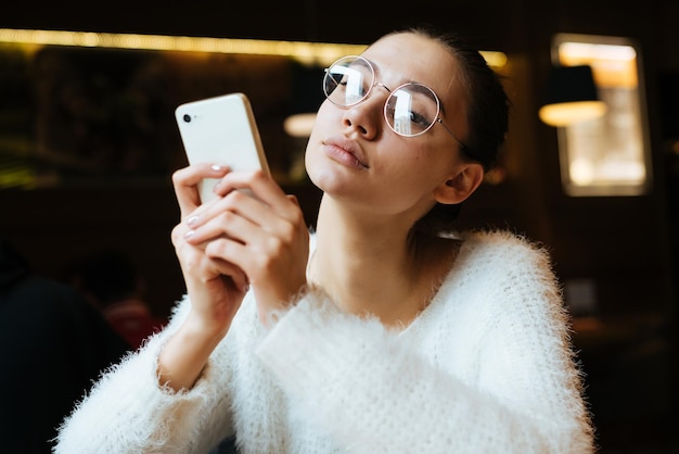 Cute young girl in glasses sits in a cafe keeps the phone and waits for her food