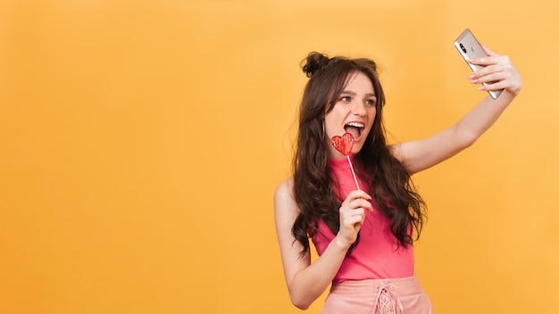 A cute young girl in pink holds a heartshaped lollipop in her hands smiles and takes a selfie on her smartphone Place for your text