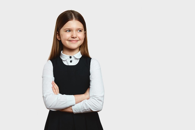 Foto ragazza carina in uniforme scolastica in piedi con le braccia incrociate e distogliendo lo sguardo nello spazio vuoto