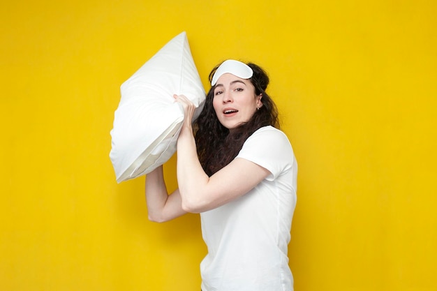 Cute young girl in sleep mask and pajamas fights with pillow on yellow background woman throws