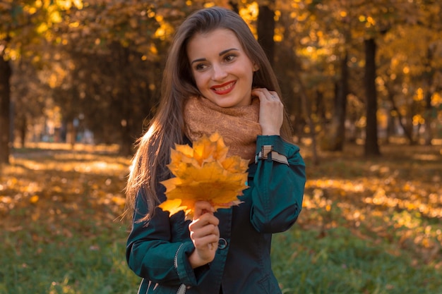 Cute young girl stands in autumn Park holding a maple leaf looks away and smiles
