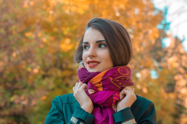 Cute young girl stands in the park looks away and keeps hands scarf closeup