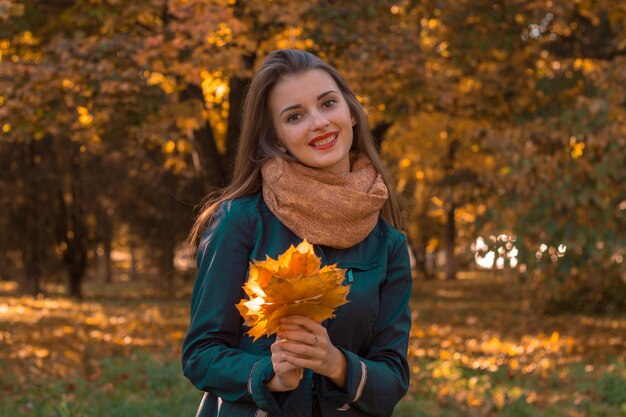 Cute young girl in a warm scarf holds the leaves looks straight and smiling