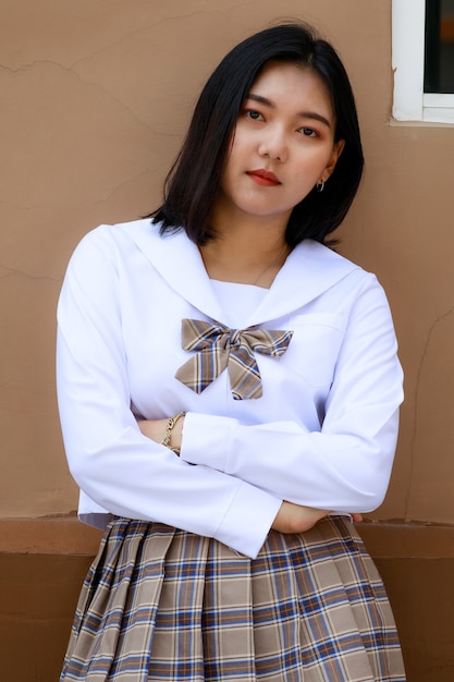 Cute and young girl wearing japanese, korean style schoolgirl\
uniform and pose to camera with fun and happy in front of school\
building.