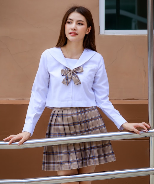 일본, 한국 스타일의 여학생 교복을 입은 귀엽고 어린 소녀가 학교 건물 앞에서 즐겁고 행복한 표정으로 카메라를 향해 포즈를 취합니다.