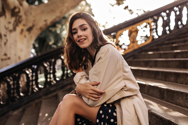 Cute young girl with brunette hair red lips beige trench coat and polka dot skirt sitting on city stairs and smiling during day walking in warm autumn city