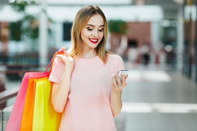 Cute young girl with light brown hair and red lips standing with colorful shopping bags, holding mobile phone, shopping concept, portrait.
