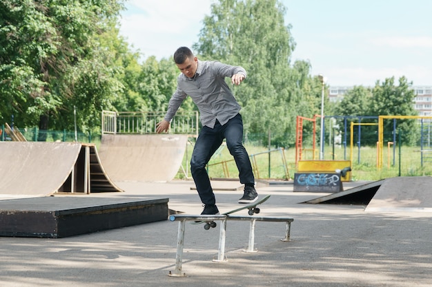 Cute young hipster guy shows trick with skateboard in skate park on warm summer day