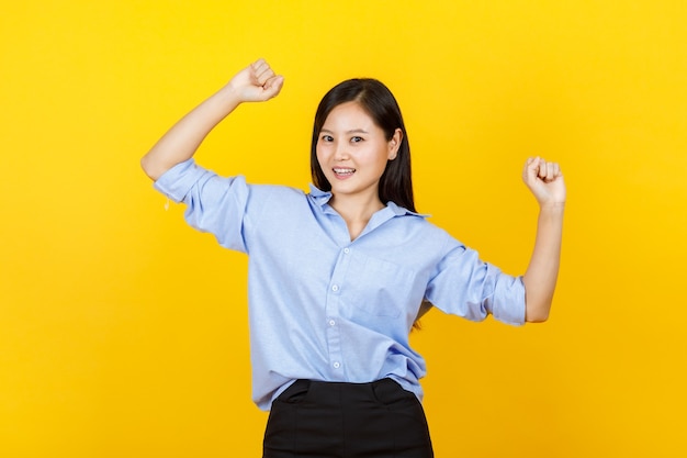 Cute and young Japanese style girl standing and raise hand show fist punches with smile face on yellow background