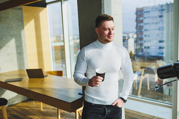 Carino giovane libero professionista maschio che beve caffè mentre è seduto in un'area di lavoro luminosa che lavora da remoto un uomo con un maglione bianco e pantaloni lavora felicemente in ufficio