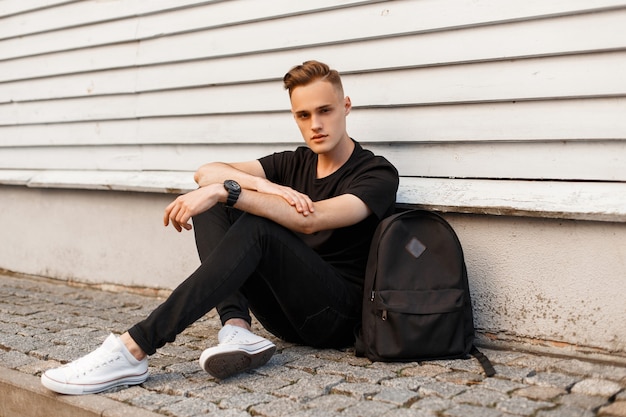 Cute young man in a black T-shirt in stylish black jeans in trendy sneakers with a black sports backpack is resting sitting on the asphalt near a white wooden vintage building