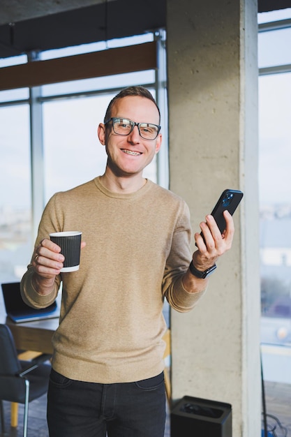Симпатичный молодой человек в коричневом свитере и очках эмоционально разговаривает по мобильному телефону и пьет кофе, отдыхая в офисе Молодой фрилансер работает удаленно