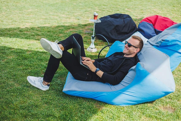 A cute young man sits in a soft chair on the street and works on a laptop relaxes and smokes a hookah Remote work