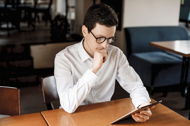 Cute young man sitting at a desk and looking at a tablet while wearing white shirt and eyeglasses.