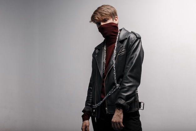cute young man wearing burgundy trendy golf in a black leather vintage jacket in black stylish jeans and metal chain, standing and posing indoors against a white wall. stylish handsome guy.