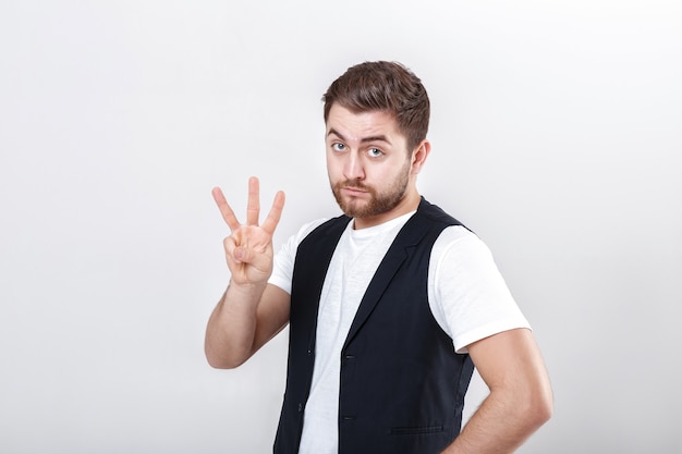 Cute young man with a beard in a white shirt and a black waistcoat showing number three gesture