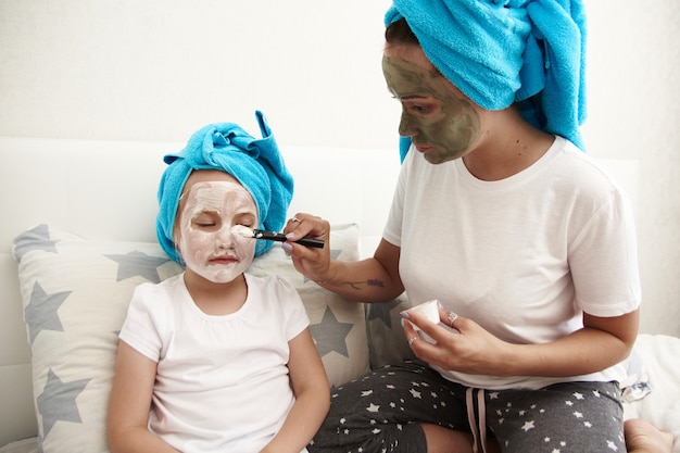 Cute young mother and her daughter are sitting on bed in towels on her head and making a face mask