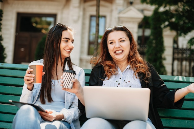 Cute young plus size women storytelling while holding a laptop on her legs and a cup of coffee in a hand while her friend is looking at her smiling.