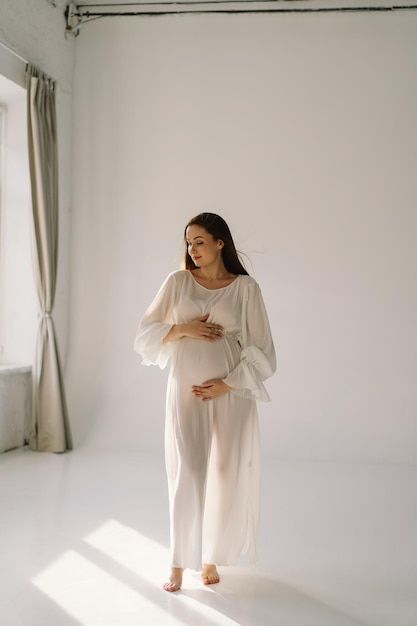 Photo cute young pregnant girl sitting on the white studio in warm light