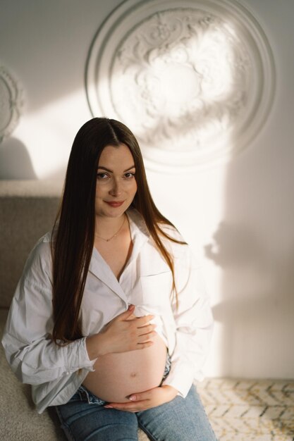 Photo cute young pregnant girl sitting on the white studio in warm light