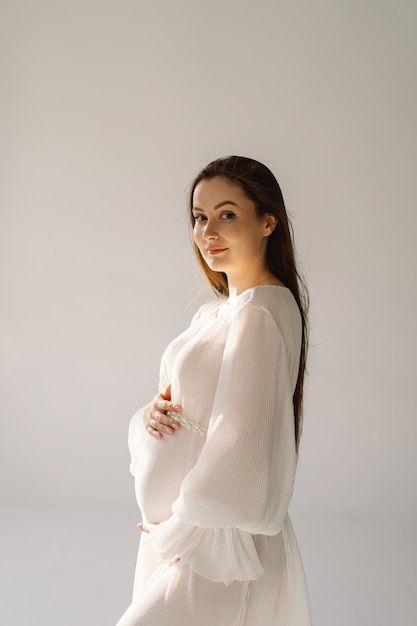 Милая молодая беременная девушка сидит в белой студии в теплом свете