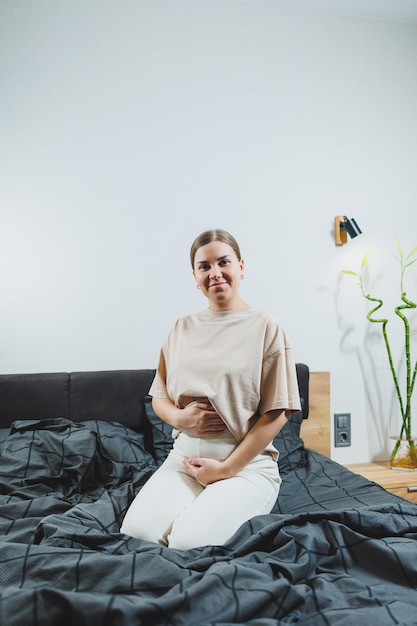 Cute young pregnant woman sitting on bed with gray sheets in casual clothes Cute pregnant woman at home