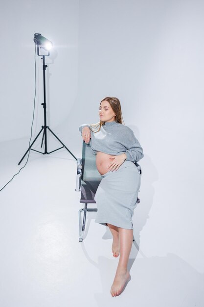 Cute young pregnant woman sitting on a chair on a white background Stylish pregnant woman in skirt posing in studio and enjoying pregnancy