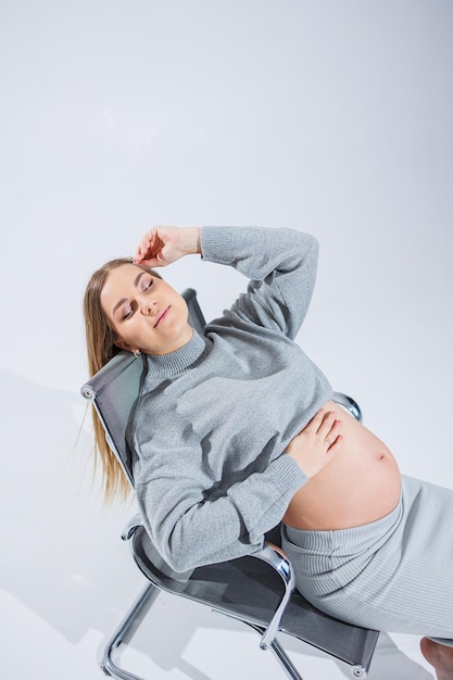 Cute young pregnant woman sitting on a chair on a white background Stylish pregnant woman in skirt posing in studio and enjoying pregnancy