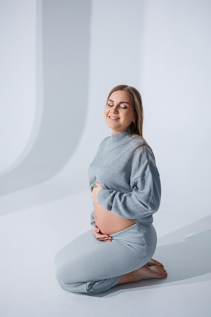 Cute young pregnant woman stroking her belly sitting on a white background Stylish pregnant woman in skirt posing in studio and enjoying pregnancy