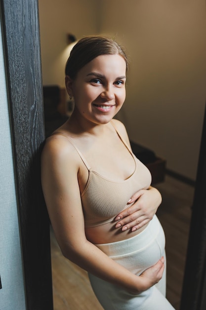 Cute young pregnant woman in a top and casual maternity clothes Cute pregnant woman hugging her belly at home