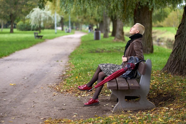 写真 赤い傘と空を見ている秋の公園のベンチに座っているかわいい若い短い髪のブロンドの女性