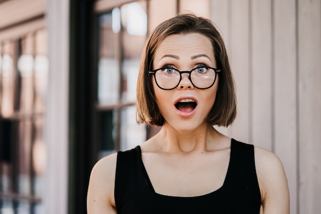 Photo cute young short haired woman  in glasses dressed casual, shocked by news with wide open eyes and mouth outdoor.