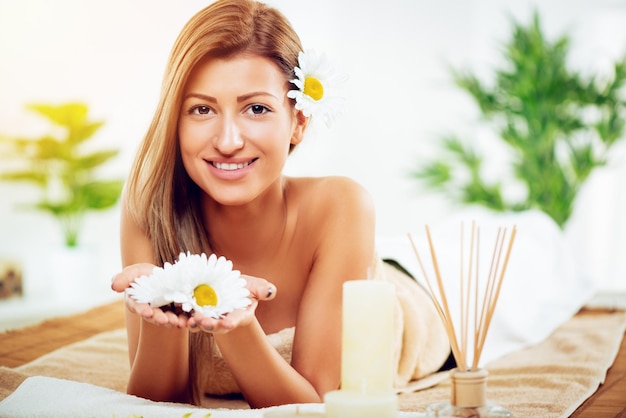 Giovane donna sorridente sveglia che gode durante un trattamento di cura della pelle ad una stazione termale. sta guardando la fotocamera e tiene in mano fiori bianchi.