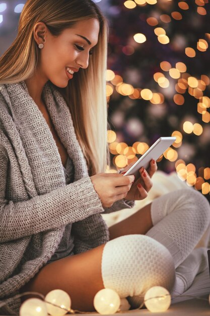 스마트폰을 사용하고 집에서 아늑한 크리스마스 휴가를 보내는 동안 웃고 있는 귀여운 젊은 여성.