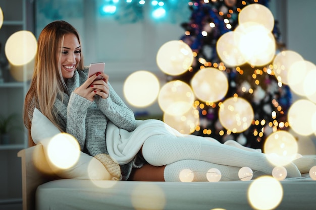 Carina giovane donna sorridente che usa lo smartphone e sorride durante le accoglienti vacanze di natale a casa.