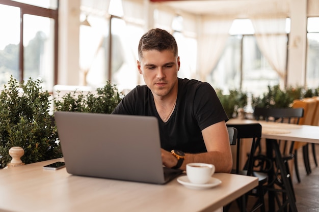 Carino giovane uomo di successo in una maglietta nera alla moda con un computer portatile si siede a un tavolo in un caffè e sta lavorando a un interessante progetto creativo su internet. ragazzo libero professionista.