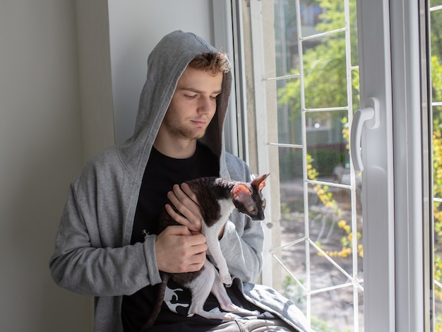 A cute young unshaven guy in a hoodie is holding a Cornish Rex cat in his arms Love for pets concept