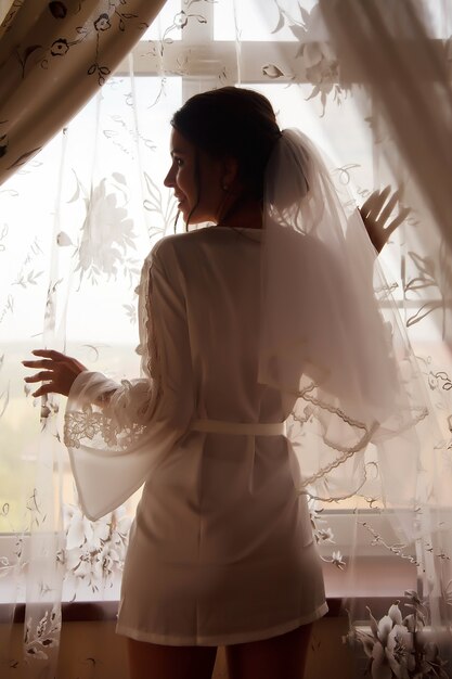 Foto sposa sveglia della giovane donna in accappatoio domestico alla finestra in casa di campagna. mattina della sposa il giorno del matrimonio. la femmina felice sta aspettando di incontrare il suo sposo. concetto di felice e lusso sposato. copia spazio per il sito