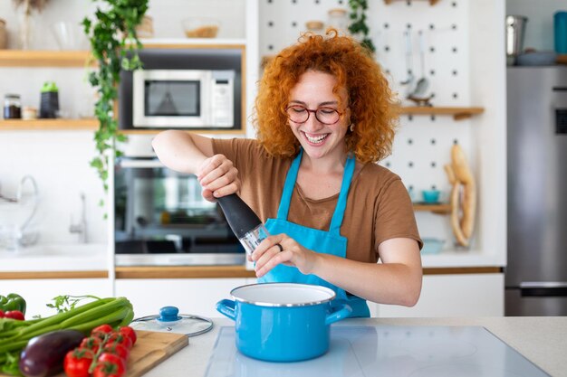 Photo cute young woman cooking and adding spice to meal laughing and spending time in the kitchen