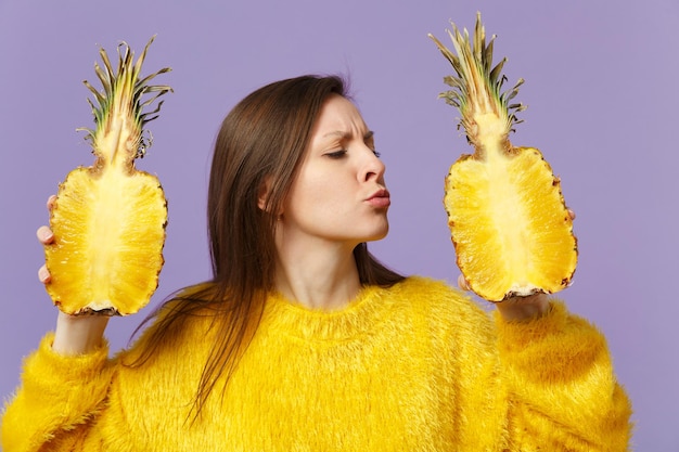 Cute young woman in fur sweater blowing sending air kiss hold halfs of fresh pineapple fruit isolated on violet pastel background. People vivid lifestyle, relax vacation concept. Mock up copy space.