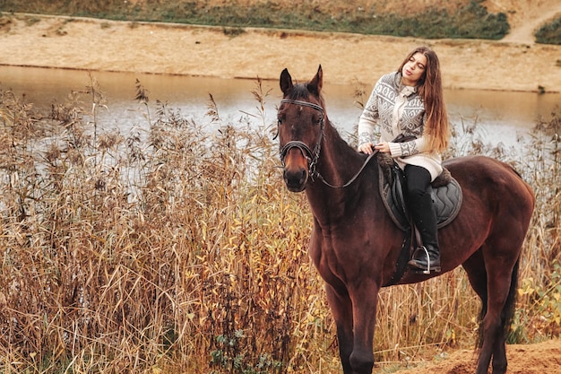 湖のそばの秋の森の馬に乗ってかわいい若い女性