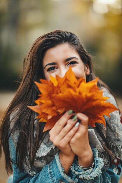 Симпатичная молодая женщина наслаждается солнечным лесом в осенних тонах. Она держит много листьев и смотрит в камеру за листьями.
