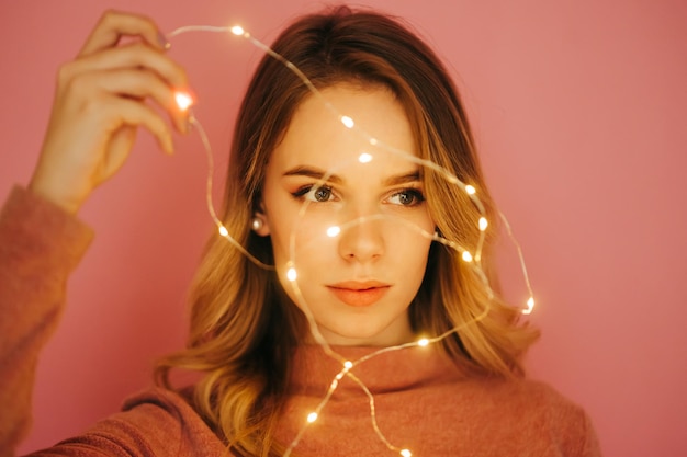 Cute young woman isolated on pink background with christmas light in hand looking aside