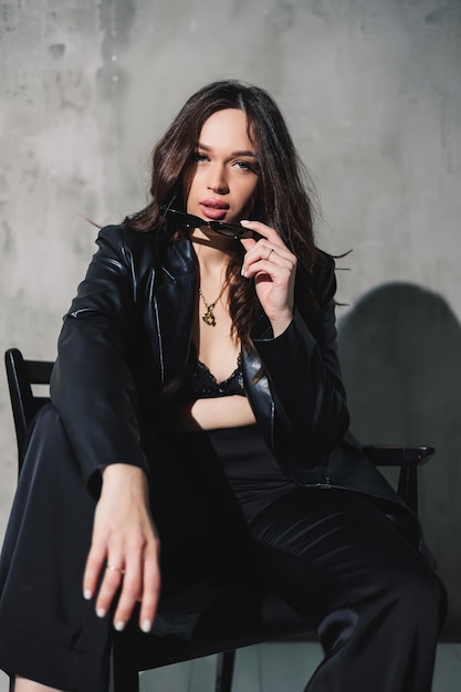 A cute young woman in a leather jacket and black pants wearing sunglasses is sitting on a chair on a gray background