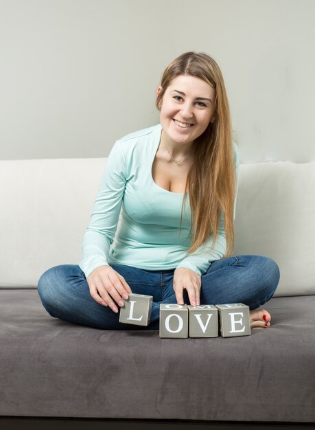 Cute young woman making word "love" from bricks on sofa